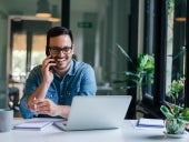 Portrait of young smiling cheerful entrepreneur in casual office making phone call while working with laptop